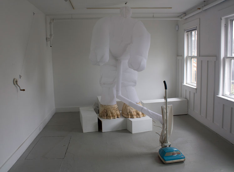 2011, Installation using vacuum cleaner on timer to inflage fabric body of figure with wooden feet. Figure can be raised or lowered by using wall crank. Figure stands on arrangement of toppled gallery pedestals.